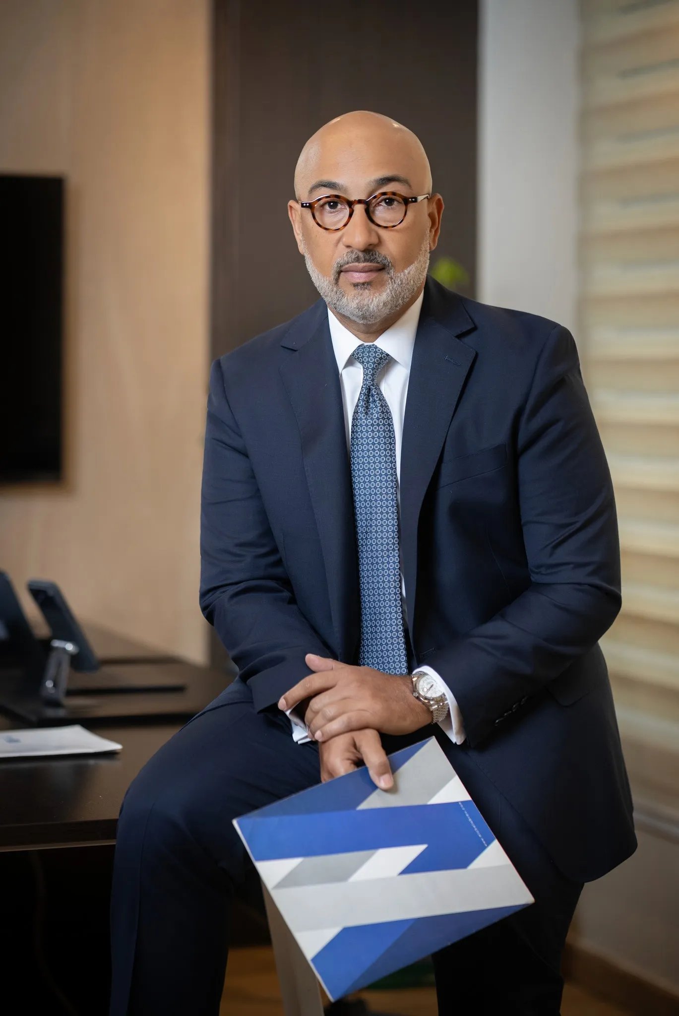 Professional headshot of a man with short brown hair and a light beard. He wears a navy blazer and gray shirt, conveying confidence and approachability.